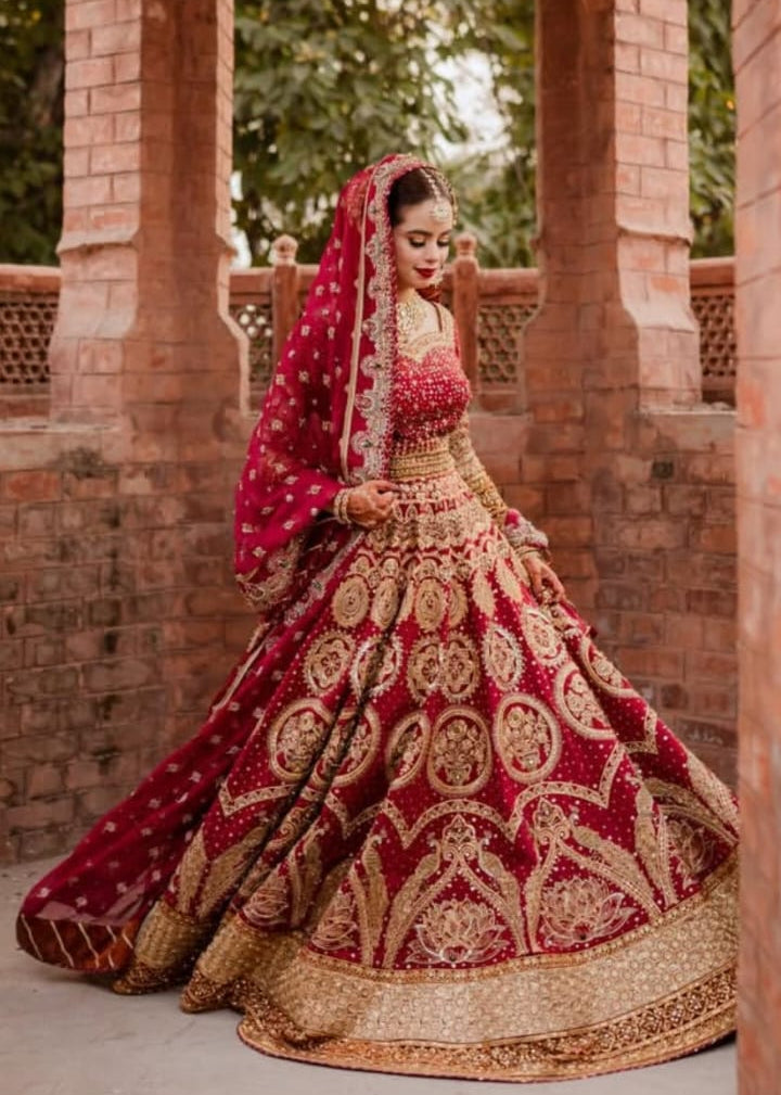 Woman in a traditional red and gold embroidered outfit standing in front of brick columns.Malika Jann by MNR
