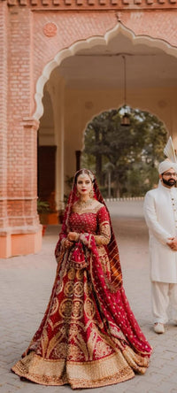 Woman in traditional red and gold bridal outfit standing in front of a man in white traditional attire, with a decorative archway in the background.Malika Jann by MNR