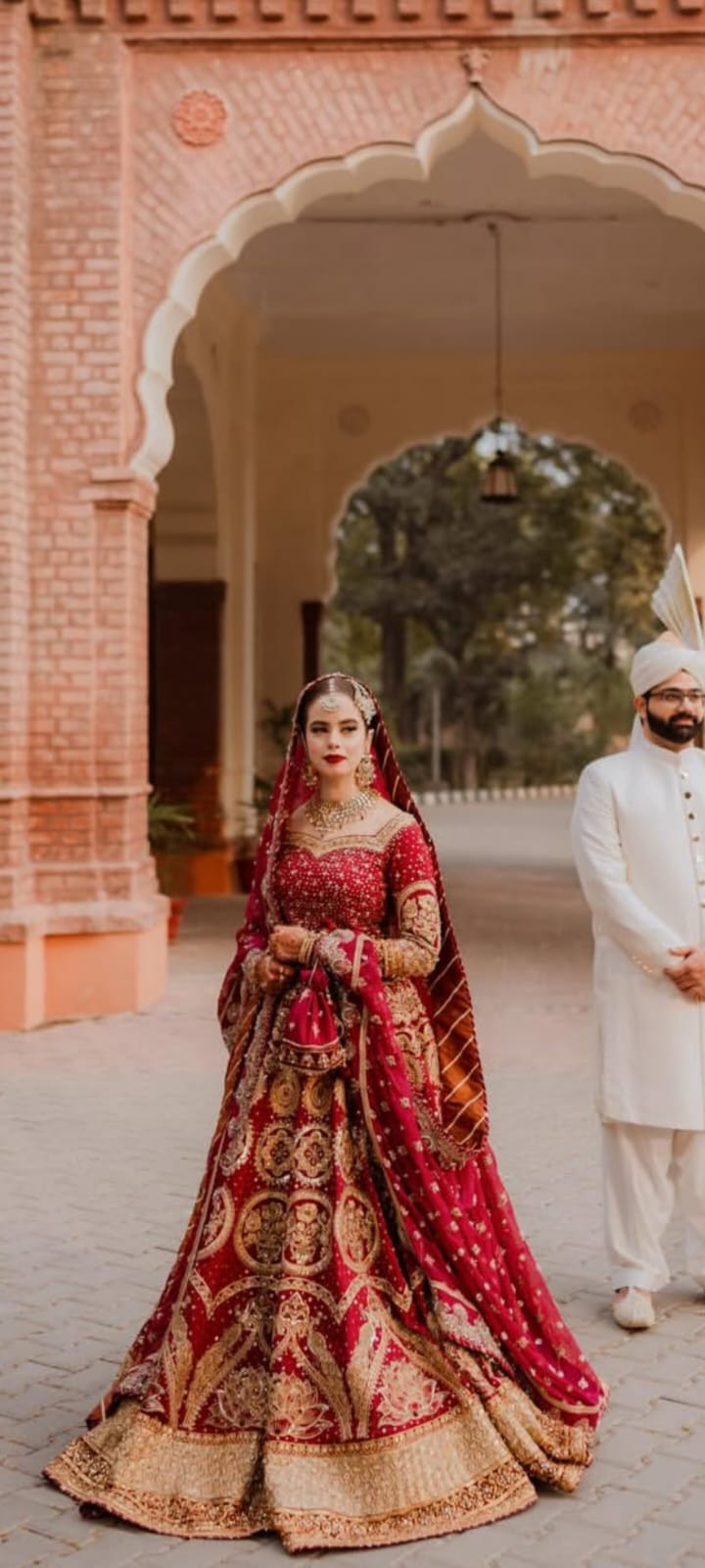 Woman in traditional red and gold bridal outfit standing in front of a man in white traditional attire, with a decorative archway in the background.Malika Jann by MNR