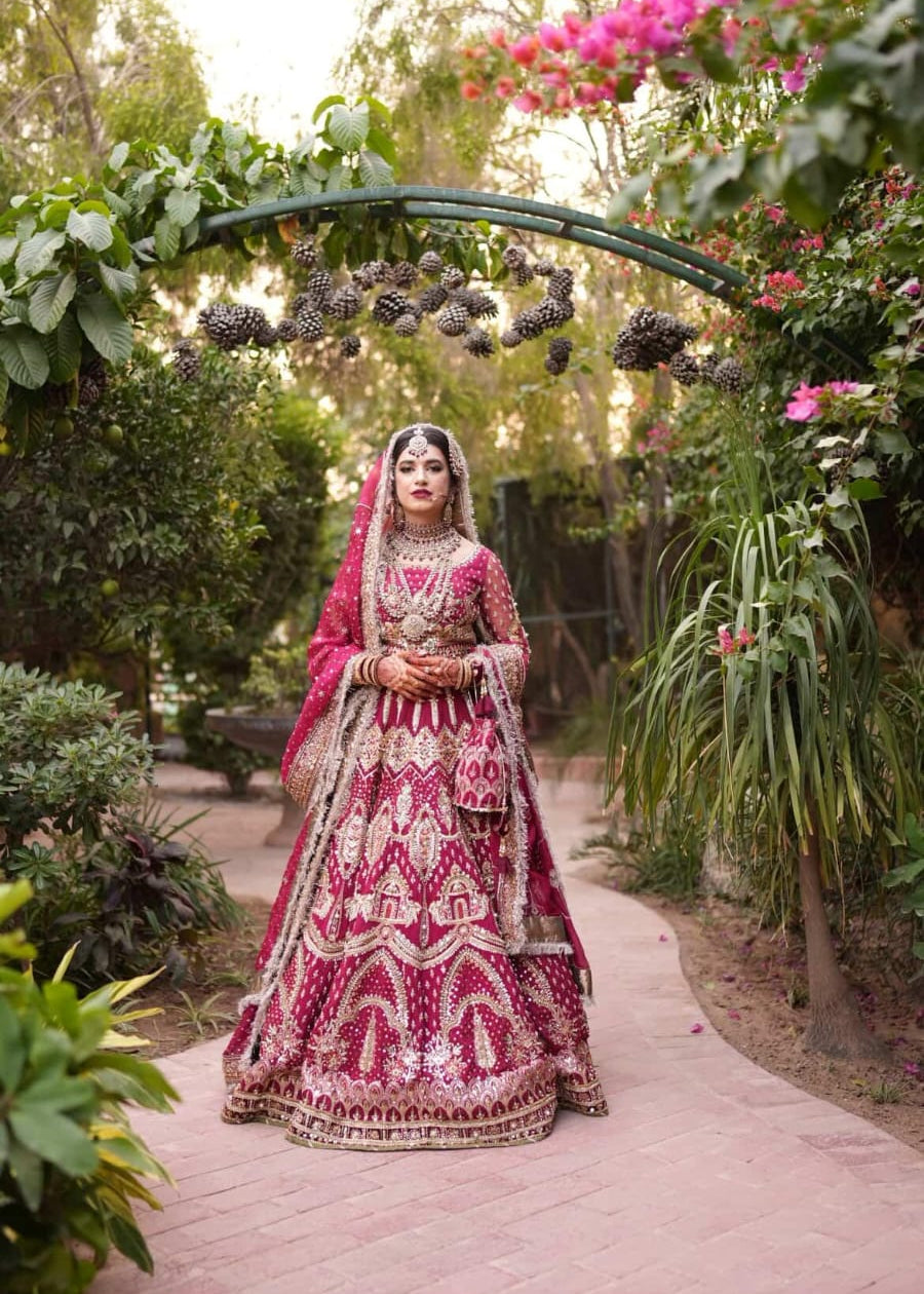 Woman in a traditional red and gold outfit standing under a floral archway in a garden.